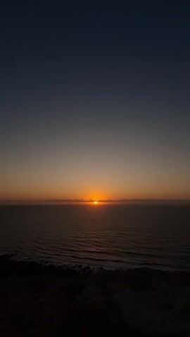 A video shows a sunset over the ocean from a cliff. The sun is setting in the distance, casting a warm orange glow across the water. The sky transitions from a deep blue at the top to a lighter orange and yellow near the horizon. The foreground of the video shows the dark silhouette of a rocky cliff. The waves are gently crashing against the shore below. The overall mood of the video is peaceful and serene.