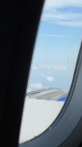 The video shows a view from an airplane window during flight. The first few seconds show the airplane's engine and wing. Then the view expands to show the wing and the ground below, with clouds in the sky. The ground appears to be a mix of green fields and some buildings. The sky is blue with white clouds.