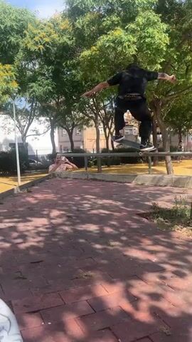 A person on a skateboard is balanced on a metal railing in a park. They are wearing a black t-shirt and black pants. The skateboard is black. The railing is silver and horizontal. Behind the skateboarder, another person is visible, lying down and looking up at the skateboarder. They are wearing a light-colored tank top. The park has trees with green leaves and yellow fallen leaves on the ground. There are buildings and a fence in the background. The ground is paved with reddish-brown bricks and sandy soil. The video cuts to the skateboarder falling off the railing and onto their skateboard, which is on the ground. They then stand up and walk away from the camera.