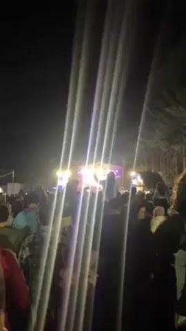 A crowd of people are gathered at a night-time concert. The stage is brightly lit with red, white and purple lights. A band is performing on stage, behind a large display of the letters "FM". Some people in the audience are recording with their phones. The lights are diffused and the light beams are visible. Trees are on the edge of the crowd and in the background.
