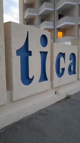 A camera pans across a sign with blue lettering on beige blocks, spelling out part of a word. The building behind the sign has balconies and orange lit windows. A swimming pool is visible past the sign.
