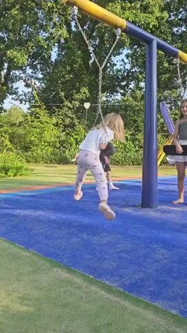 A blond-haired child is swinging on a swing. They are wearing a white shirt and pink patterned pants. The child is in mid-swing, with their legs extended and hair flying. In the background, another child, a boy, stands on the blue ground, and a girl stands near a purple swing set. The swing set is blue and yellow, with metal chains. The ground is a blue rubberized surface with green grass on the left. Trees are visible in the background.