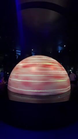 The video shows a large, hemispherical dome-shaped object illuminated from within. The exterior of the dome displays a swirling, marbled pattern of pink and white. Thin, horizontal lines of light are visible across the surface of the dome, moving from left to right. The dome is positioned in a dimly lit, circular room. In the background, out of focus, several people are visible, some standing and observing the dome. The overall impression is that of a scientific or exhibition display.