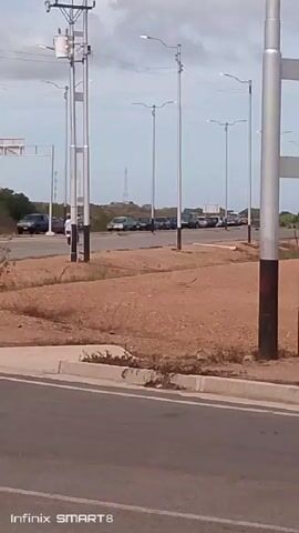 The video shows an outdoor scene on a partly cloudy day. A road runs across the bottom of the frame, with a low concrete barrier separating it from a patch of dry, brown land. Beyond this area is another road with several parked vehicles, including cars and SUVs. A person on a motorcycle is driving on the further road, moving from left to right in the frame. Power lines and tall lampposts are visible along the roadsides. In the background, an antenna tower is visible. The video is captured by an Infinix SMART 8 phone, as indicated in the lower left corner.