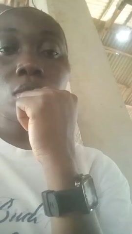 The video is a close-up shot of a dark-skinned person wearing a white t-shirt with some visible writing. The person has a neutral expression and is resting their chin on their fist. They are wearing a watch on their left wrist with a black strap and a square face. The background appears to be indoors, with a wall and a metal roof structure visible. The lighting is somewhat dim.