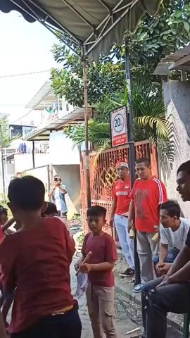 The video shows an outdoor gathering of people, likely a community event or party, with a focus on children. In the foreground, a person wearing a denim jacket faces away from the camera, holding a microphone, suggesting they are an organizer or performer. Several children in red shirts are visible, some appearing engaged in an activity or performance, possibly a game or dance. In the background, other adults and children are present. There are decorative items and boxes on the ground, implying gifts or prizes. The environment appears to be a street or alleyway with residential buildings and trees. The overall atmosphere seems festive and lively. A speed limit sign of 20 is visible on a pole.