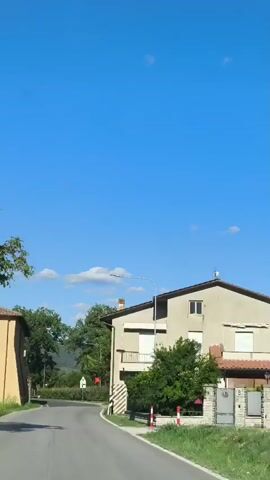 The video shows a view from a moving vehicle looking forward onto a paved road that curves slightly to the right. On the left side of the road is a two-story building with a tan facade and a brown tiled roof. A large tree with green foliage is also visible to the left of the road. On the right side of the road, there is another building with a white facade and a light-colored roof. Further down the road, trees and vegetation line the sides, and there are signs visible, including a red sign and a white sign with black markings. The sky is a clear, bright blue with a few small white clouds. The overall scene suggests a rural or semi-rural setting on a sunny day.