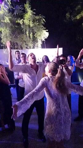 A group of people are dancing at an outdoor nighttime event. In the foreground, a man in a white shirt and black pants and a woman in a sparkly white dress are dancing facing each other. The man has his arms raised and is smiling, while the woman has her arms extended. Other people are visible in the background, some of whom are also dancing or watching. The environment appears to be a courtyard or patio with trees and white structures. Blue and white lights are illuminating the dance floor.