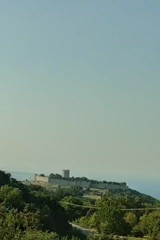The video shows a landscape from a moving vehicle, likely a car, as evidenced by the motion blur of the trees in the foreground. The sky is a clear, pale blue. In the distance, on a hilltop, a stone structure, possibly a castle or fortress with crenellated walls and a tower, is visible. The surrounding terrain is green and hilly, with trees and vegetation covering the slopes. There are glimpses of the sea in the far distance. The overall impression is of a sunny day in a rural, possibly Mediterranean, setting. The audio consists of indistinct speech in what sounds like a Slavic language, possibly Polish or a similar dialect, with a woman's voice speaking. The speech is not clearly understandable.