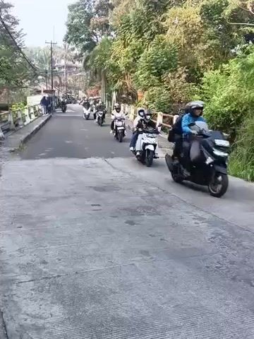A group of motorcyclists are riding down a paved road. The road is lined with trees and a white railing on the right side. There are buildings visible on the left side of the road in the background. Some of the motorcyclists are wearing helmets. One motorcycle in the foreground is carrying two people. The road appears to be slightly uphill.
