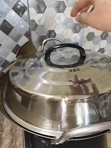 The video begins with a close-up of a stainless steel pot with a lid. A hand is seen lifting the lid. The lid is then completely removed, revealing the contents of the pot. The pot contains a dish with chicken and potato wedges in a curry sauce. The curry sauce is a rich, yellowish-orange color. The chicken pieces appear to be small and dark brown, while the potato wedges are large and pale yellow. The pot is a wide, shallow wok-like pan with two handles. The background consists of a tiled wall with a geometric pattern of gray, black, and white hexagons. The scene appears to be a kitchen environment.