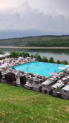 The video shows a wide outdoor swimming pool with a vibrant blue interior. Many people are swimming and relaxing in the pool. Surrounding the pool is a deck area furnished with numerous lounge chairs and umbrellas, many of which are empty. To the left of the pool area, there is an inflatable water slide and several white tents that may be used for seating or dining. In the background, a calm body of water, likely a river or lake, stretches out, bordered by a tree-covered hill. The sky appears overcast, with clouds suggesting the possibility of rain. The foreground features a grassy slope.