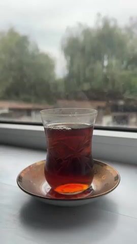 A close-up shot focuses on a clear glass of dark tea with steam rising from it, resting on a decorative copper saucer. The saucer is placed on a white windowsill, with a blurred background of trees and a building visible through a window. The lighting is soft and overcast, suggesting an outdoor or ambient light source. The video is still with no discernible movement.