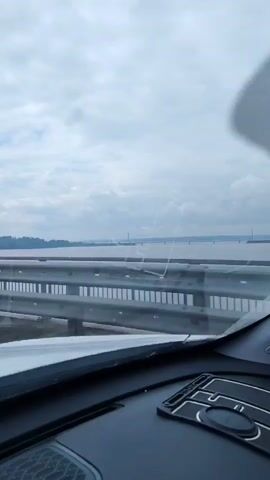 The video shows a car driving over a bridge. There is a wide river or body of water under the bridge. The sky is cloudy. The car's dashboard is visible. A segment of metal guardrail is visible on the side of the bridge. In the distance, the shore is visible along with some buildings and power lines.