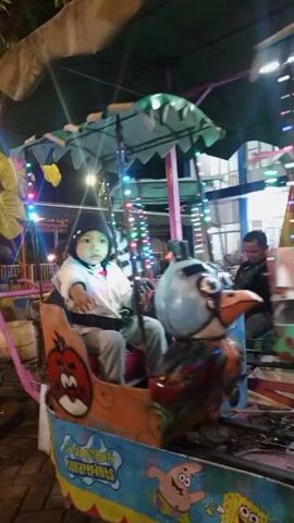 The video shows a child in a small amusement park ride. The child is wearing a black beanie and a light-colored jacket over grey pants and is sitting in a boat-shaped car. The car has cartoonish drawings on its sides, including an angry red apple and a green bird. The ride appears to be circular and is decorated with colorful lights. In the background, other amusement park rides and people can be seen. The lighting suggests it is nighttime or indoors in a dimly lit area. The child is looking forward and seems to be holding onto a steering wheel-like object.