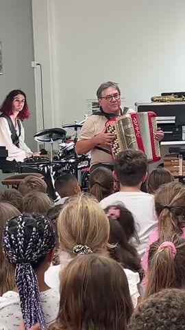 A man plays an accordion in front of a group of children. He is wearing glasses and a t-shirt. To his left, another person plays a drum set. The children are seated on the floor, facing the musicians. The environment appears to be an indoor space, possibly a classroom or auditorium.