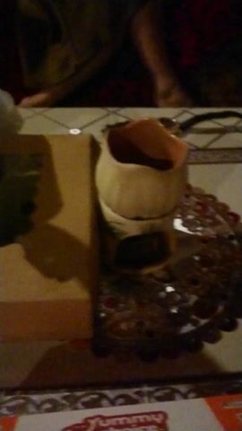 The video begins with a close-up, slightly out-of-focus shot of what appears to be the top edge of a light brown cardboard box, with a white, possibly textured, element behind it. A white plastic bag is partially visible on the upper left.

As the camera slowly pans downwards and to the left, more details come into view. A brown cardboard box with a white circular sticker, possibly a logo or label, is visible on the right side of the frame. Below it, resting on a patterned surface, is a bright green plate or shallow bowl.

Inside this green plate, several baked goods are visible, each individually wrapped in clear plastic. These items appear to be golden-brown and somewhat round or oval in shape, resembling bread rolls or pastries. Some of them exhibit an orangish-red hue, possibly from a topping or a type of bread. A white plastic bag is also visible on the far left, partially obscuring some of the items.

The background on the left consists of a dark red fabric with a subtle pattern, while the surface on the right has a lighter, possibly tiled or checkerboard pattern, with a shiny, decorative border on the extreme right. The lighting is dim, suggesting an indoor setting with artificial light. The camera holds steady on the green plate with the wrapped items for the remainder of the video.