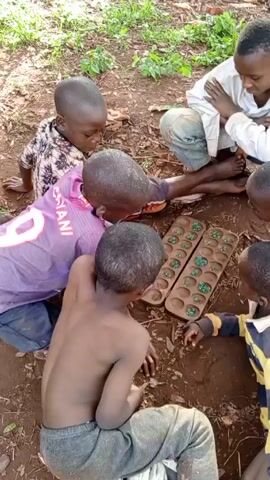 The video shows a group of five young boys playing a traditional board game on the ground. The game board is made of wood, has two rows of six or seven indentations each, and contains green marbles or stones within these indentations.  The boys are sitting or kneeling around the game board on bare ground in an outdoor setting. Patches of grass and small green plants are visible in the background. The boys' clothing includes a white button-down shirt, a purple soccer jersey, and a striped yellow-and-black shirt. The video is shot from an elevated perspective.