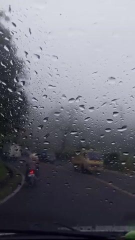 A stationary view from inside a car shows a rain-streaked windshield with raindrops obscuring the view. Through the windshield, a road is visible, winding slightly to the left.  A motorcycle with a single rider and lit taillight is traveling on the road in the same direction as the viewer. Other vehicles, including cars and trucks, are also present on the road. The weather is foggy and overcast, with trees lining the side of the road. The overall environment suggests a drive in a misty, rainy, mountainous area.