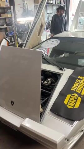 The video shows a white Lamborghini with its gull-wing doors open. The front hood is also open. A black NAPA Tools & Equipment mat sits on the front of the car, near the open hood. The Lamborghini emblem is visible on the hood.  Three men are visible in the background, one with a baseball cap and beard, another with a tattoo on his neck.  The setting appears to be an automotive repair shop.