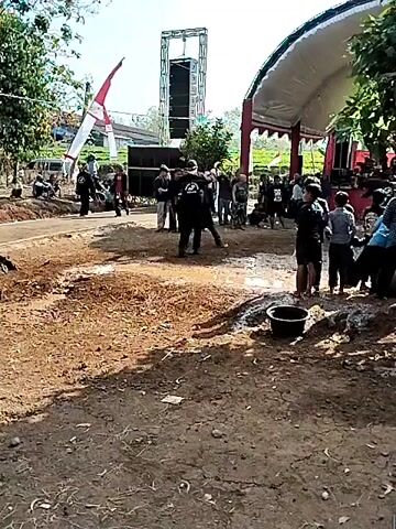 The video shows an outdoor gathering. In the foreground, there is disturbed earth and some puddles of water. In the middle ground, a group of people are gathered, some standing and some sitting. There is a stage covered with a white tent and decorated with red trim. Behind the stage, there are large speakers and what appears to be a tall lighting rig. There is also a flag with red and white stripes visible in the background. Two individuals in the middle ground are dressed in traditional attire that includes elaborate headpieces and patterned sashes. They appear to be performing or reenacting a scene, with one individual interacting with a large, animal-like costume. Other people in the crowd are watching the performance. The overall atmosphere appears to be festive, possibly a cultural event or celebration.