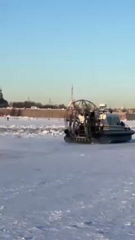 The video shows a hovercraft moving across a snowy landscape, kicking up a large cloud of snow dust. In the background is a cityscape with prominent architectural details, including what appears to be a tall, golden spire and other building structures with domes. Several people are visible on the frozen surface, near the base of the distant building. The sky is clear and blue. The lighting suggests it is daytime, likely late morning or early afternoon, casting bright reflections on the snow. The hovercraft is equipped with a large, circular fan on the top.