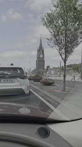 A video recorded from inside a vehicle shows a clock tower in the distance. The vehicle is in traffic on a street with other cars. Flower planters are visible on the median. The clock tower is a prominent structure with a pointed spire and clock faces. The sky is cloudy.