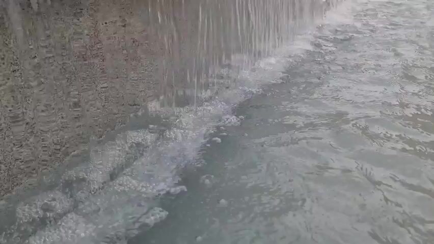 This is a close-up, low-angle video of a man-made waterfall, likely a decorative feature in an outdoor setting. The water cascades down a textured, grey wall on the left side of the frame, creating a foamy, churning effect as it hits the pool below. The water in the pool is a pale blue-grey color, and small ripples and waves are visible. The lighting suggests it could be daytime, but the exact time of day or weather conditions are not discernible from the footage. There is no visible text in the video. The scene evokes a sense of tranquility and the natural beauty of flowing water. Given the location context of Plungė, Lithuania, this waterfall could be part of a park, garden, or public space within the town.