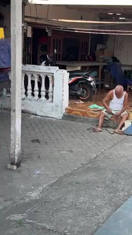 An elderly man with a shaved head and wearing a white tank top and light-colored pants sits on the ground in front of a building. He is facing to his right and appears to be working on something in his lap with his hands. To his left, there is a concrete pillar. To his right, there is a gray paved walkway and a tiled area in front of the building. In the background, a white motorcycle is parked. The building has a covered entrance with large glass doors and a white railing. Blue tarps are visible in front of the building. The ground is a mixture of brick paving and concrete.