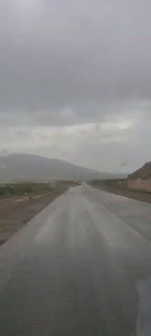 The video captures a first-person perspective from inside a vehicle, looking straight down a wet, asphalt road. The sky is heavily overcast and grey, with distant mountains or hills appearing hazy and muted under the thick cloud cover. The landscape alongside the road is arid and sparsely vegetated, featuring dry earth and scattered scrub.

Around the 0:03 mark, the view shifts slightly to prominently display numerous raindrops adhering to the windshield, blurring the distant scenery and highlighting the ongoing rain. The wet road surface reflects the dull light from the sky. Far in the distance, another vehicle can be faintly seen ahead, moving along the straight road. The scene suggests a rural or remote setting, consistent with the provided location context of Kan Mehtarzai, Pakistan, during a rainy or inclement weather period. No people are visible, and there is no discernible text in the video. Background music is playing throughout the clip.