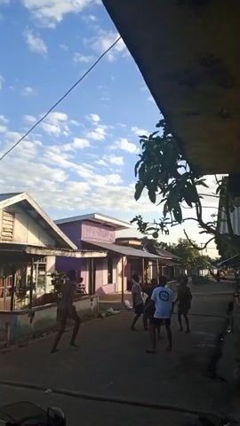 A group of young males are playing a game of volleyball in a street in front of houses. The sky is blue with scattered white clouds, and the sun is shining. The houses are simple, some with wooden elements and some painted in bright colors. The street is paved and appears to be a residential area. The players are dressed casually in shorts and t-shirts. Their actions suggest an energetic game, with some players jumping and reaching for the ball.