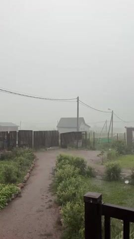 The video shows a cloudy and rainy day in a rural area. The foreground features a dark railing and some greenery. A dirt path leads into the scene, lined with plants and a wooden fence. In the background, there are several houses, a small shed, and utility poles with wires. The overall atmosphere is hazy due to the rain.