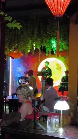 The video shows a band performing live music in what appears to be a restaurant or bar. The band consists of five members: a singer, a guitarist, a keyboard player, and what appears to be a percussionist. They are performing on a small stage in front of a backdrop that is painted with what looks like a colorful cosmic scene with a bright, central moon.

Several customers are seated at tables watching the performance. In the foreground, a woman with blonde hair is seated at a table with a camera on a tripod, presumably recording the performance. There's also a man in a white shirt sitting at the same table. A table lamp and two glasses are on the table.

The venue is decorated with hanging greenery and red lanterns, creating a warm and inviting atmosphere. The lighting is dim, with colored lights illuminating the stage and the band.