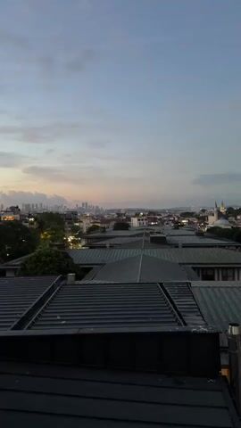 A vertical video shows a city skyline at dusk. In the foreground, the dark green, ribbed roofs of modern buildings are visible. Beyond these, a sprawling cityscape stretches into the distance, characterized by a mix of red-tiled roofs and other structures. Prominently featured in the middle ground is a large domed building with minarets, likely a mosque or historical structure, and a tall, slender white minaret stands to its right. In the far distance, a cluster of modern skyscrapers forms a faint outline against the horizon. The sky above is a gradient of pale blue transitioning to a soft pinkish-orange near the horizon, with scattered clouds. The overall impression is a view of a city with historical and modern elements under a twilight sky. There are no people or discernible actions taking place.