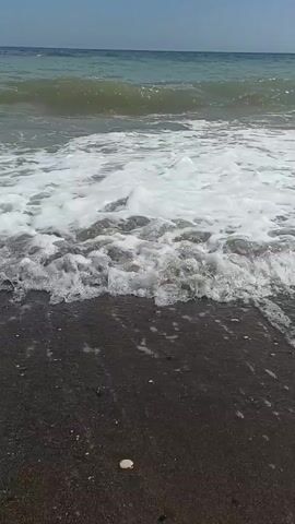 This video depicts a beach scene with a focus on the ocean and shoreline. The water is a light blue-green with white foam from waves crashing onto the shore. The beach is covered in dark sand and small pebbles, with a few scattered shells. The sky is bright blue with scattered white clouds. There are several people visible in and near the water. In the foreground, a person is sitting on the wet sand with waves washing around them. Further out in the water, a few other people are swimming and playing in the waves. In the distance, along the shoreline, there is a line of trees and buildings, suggesting a populated area. The overall atmosphere is one of a sunny day at the beach.