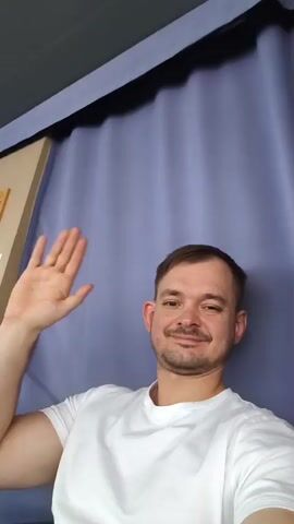The video begins with a medium shot of a man wearing a white t-shirt. He is smiling and waving to the camera. He has short brown hair and a light beard. He is in front of a purple curtain and a beige partition. The camera then pans up and to the right, showing a glimpse of the ceiling and a blurred view of what appears to be an interior space with fluorescent lights. The camera then moves to a different scene, showing a radar display screen. The screen shows a circular radar image with various targets and information. The screen is positioned on a console with other equipment and displays. Large windows with white frames are visible in the background, suggesting an indoor location with natural light.