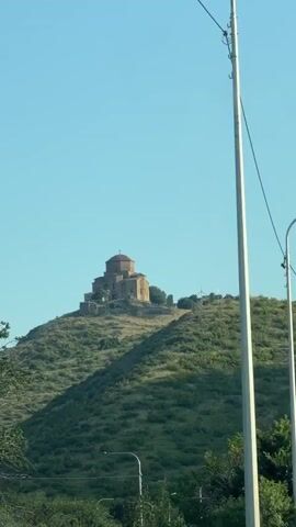 This is a short video clip of a church on a hilltop. The camera appears to be moving, possibly in a vehicle, as indicated by the slight camera shake and the presence of utility poles and wires in the foreground. The church is made of stone and has a dome. The hilltop is covered in green vegetation. The sky is clear and blue. The video is very short, lasting only a few seconds.