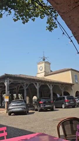 The video shows an outdoor market with a covered wooden structure. There are several cars parked in the area, including a red Renault Clio, a silver Ford Puma, a black Renault Kadjar, and a grey minivan. The market has a clock tower with a beige facade. In the background, a church steeple is visible. The sky is clear and blue, and there are some green leaves in the foreground, suggesting it is daytime.