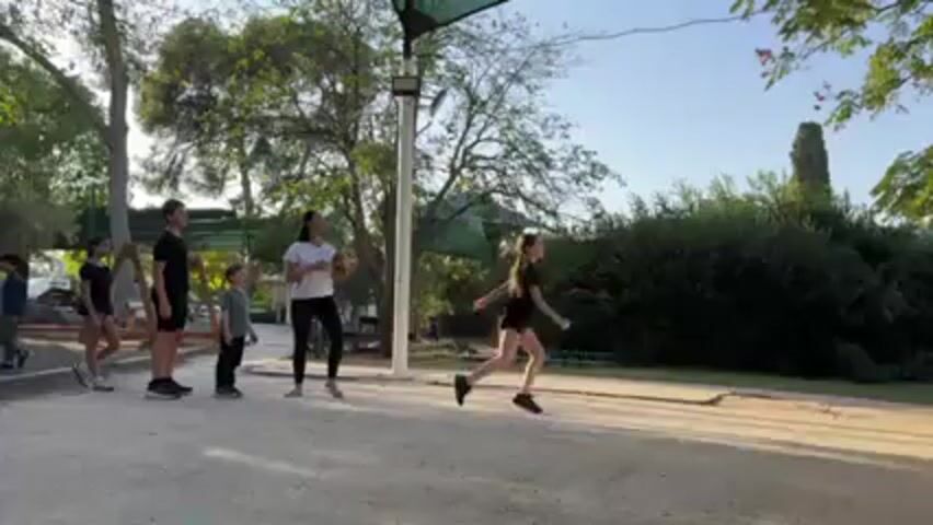 The video shows a group of five people, likely a family, in an outdoor park setting. A woman in a white shirt and black leggings throws a basketball to a young girl in black athletic wear. The girl catches the ball mid-jump. To the left, stand two other children, a teen, and a young boy who are observing the activity. They are all facing the woman and girl. The setting appears to be a park with trees, some playground equipment visible in the background, and a gravel path. The sky is clear, suggesting a sunny day.