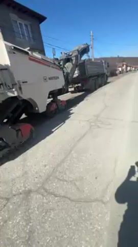 The video shows a Wirtgen asphalt milling machine removing asphalt from a street. The machine is white and gray, with red accents, and has tracks for movement. It is positioned on a paved street in a residential area, with a building visible in the background. The milling machine is loading the asphalt it removes into a dark-colored dump truck driving in front of it. The sky is blue, and it appears to be a sunny day. A person holding the camera is partially visible on the right side of the video.