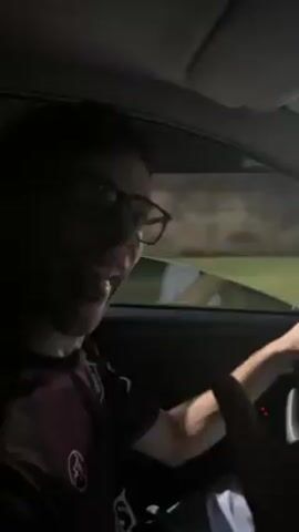 The video shows a person driving a car at night. The person is wearing glasses and has dark hair. The car is driving on a street with streetlights. The video is shot from the perspective of the driver. The dashboard is visible. There are stickers on the windshield. Trees line the street.