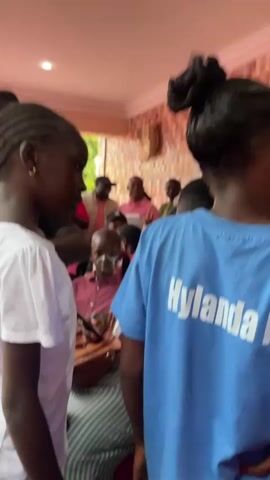The video shows a group of people, primarily children and adults, gathered outdoors. The focus is on several young girls, two of whom are in the foreground. One girl in the left foreground is wearing a white t-shirt and has braided hair. Another girl in the right foreground is wearing a blue t-shirt with the text "Hylandai" visible on the back. A young boy with face paint is also visible in the lower left foreground. In the background, a woman in a floral top with glasses is speaking and holding a phone. Other adults and children are present, some are looking in various directions, and there is an indication of an outdoor structure with columns. The overall atmosphere appears to be a gathering or event.