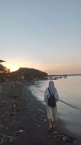 A person wearing a hijab and a backpack walks along a beach at dusk. The beach is covered in dark sand and debris. In the distance, there are some wooden structures and boats. The sun is setting over the ocean, casting a warm glow on the sky. The waves are gently lapping at the shore. The person is walking away from the camera, towards the ocean.