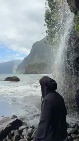 A man in a black hooded sweatshirt stands on a rocky beach near a waterfall that empties directly into the ocean. The waterfall is on the right side of the frame, partially obscured by foliage. The ocean stretches out to a hazy mountainous coastline in the distance. The sky is partly cloudy. The man initially faces away from the camera, then turns, smiles, and stretches out his arms. He then turns away again.