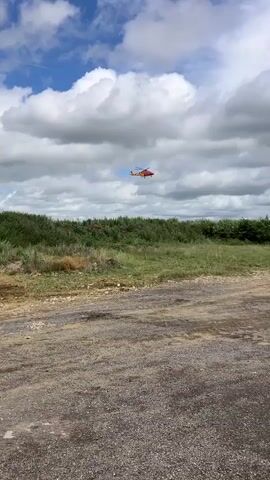 A red and yellow helicopter flies across a cloudy blue sky above a field of green grass and trees. The helicopter appears to be leaving a trail of smoke or vapor behind it. In the foreground, there is a dirt and gravel path. In the background, a large white tanker truck is parked on the right side of the frame.