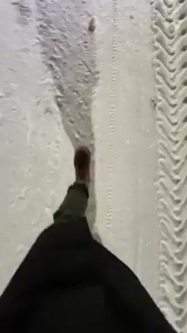 The video shows a person's view while walking on a snow-covered surface. The person is wearing brown boots, dark pants, and a dark coat. A tire track is visible on the right side of the frame. The snow is packed and uneven. The person's shadow is visible on the snow.