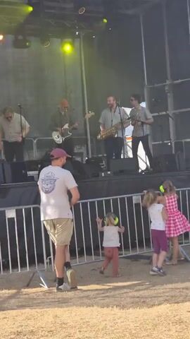 A live band performs on an outdoor stage. The band consists of at least four musicians, including a guitarist, a saxophonist, and a singer. They are positioned on a raised stage with a black backdrop and overhead lighting. In front of the stage, there is a metal barricade. Behind the barricade, two young girls are visible. One girl, wearing a pink and white polka dot dress and purple leggings, is dancing. The other girl, in a pink dress and white top, is also dancing. A man in a light-colored t-shirt and shorts stands to the left, facing the stage. The ground appears to be covered in sand or dirt. The overall scene suggests an outdoor concert or festival.