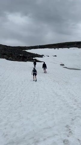 Three people walk up a snow-covered hill on a cloudy day. The person in the lead is wearing dark shorts and a light shirt, carrying something white in their right hand. The second person is wearing a dark jacket and dark pants. The third person is wearing a dark top and dark shorts. A fourth person with red hair, wearing a dark top, appears in the bottom left corner, holding a walking stick and looking to the left. In the background, there's a rocky hill, and the sky is overcast.