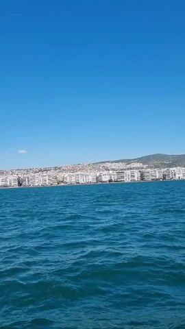 A low-angle, vertical video shot from a boat shows a blue sky and choppy blue water. In the distance, a densely populated coastal city with white buildings stretches along the shoreline. A verdant hill rises behind the city. The video is approximately 5 seconds long.