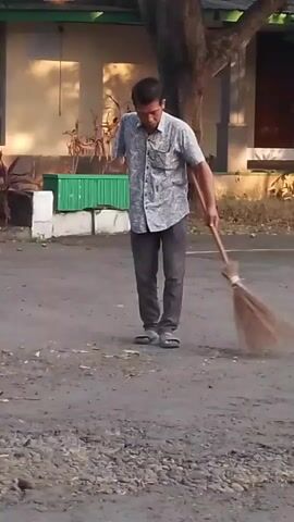 A man with one arm is sweeping the ground with a broom. He is wearing a patterned short-sleeved shirt and dark pants. The ground is dusty and uneven, with some small stones. In the background, there is a building with windows, some plants, and a tree. The man pauses sweeping to listen to something or someone on his phone, which he is holding to his ear with his right hand.